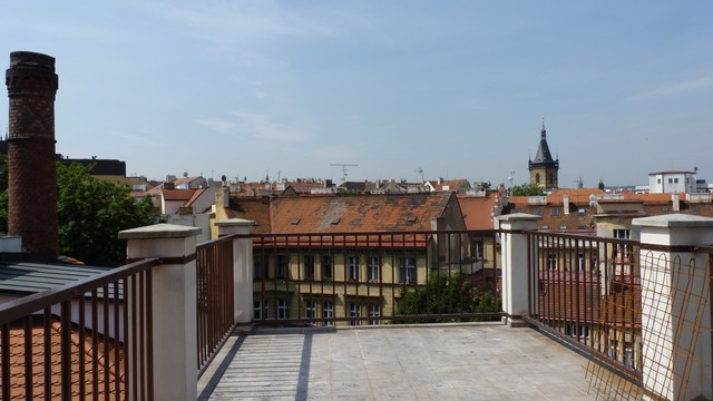 Pohled z terasy půdního bytu ukazuje rozlehlou panorama střech pražských domů a věží, v popředí terasa s elegantním zábradlím, v pozadí panorama Prahy.