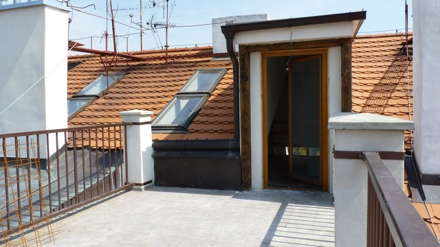 Prostorná terasa půdního bytu s dřevěnými dveřmi a okny, obklopená typickou pražskou střechou s taškami.